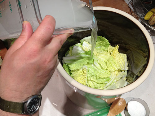 brine being poured over nappa cabbage leaves in a fermentation crock 