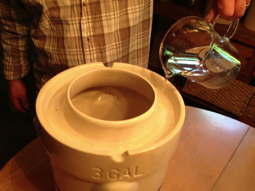 water being poured into the rim of the pickling crock to seal out air 